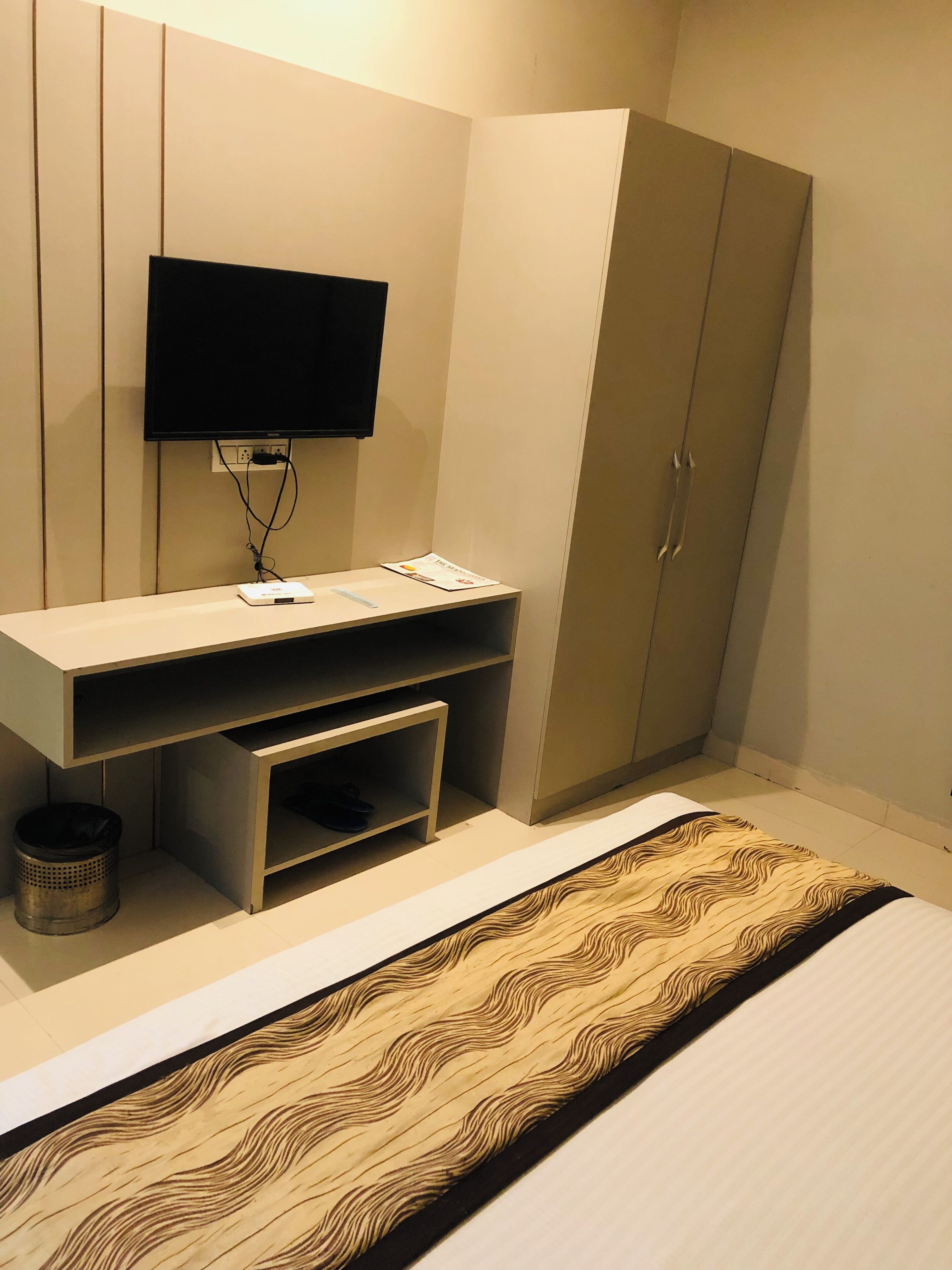 A bedroom with a bed, a TV mounted on the wall, a wardrobe, and a desk with a computer and some items on it.