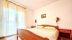 A bedroom with a wooden bed, a nightstand, a lamp, a window with curtains, and a wall-mounted picture.