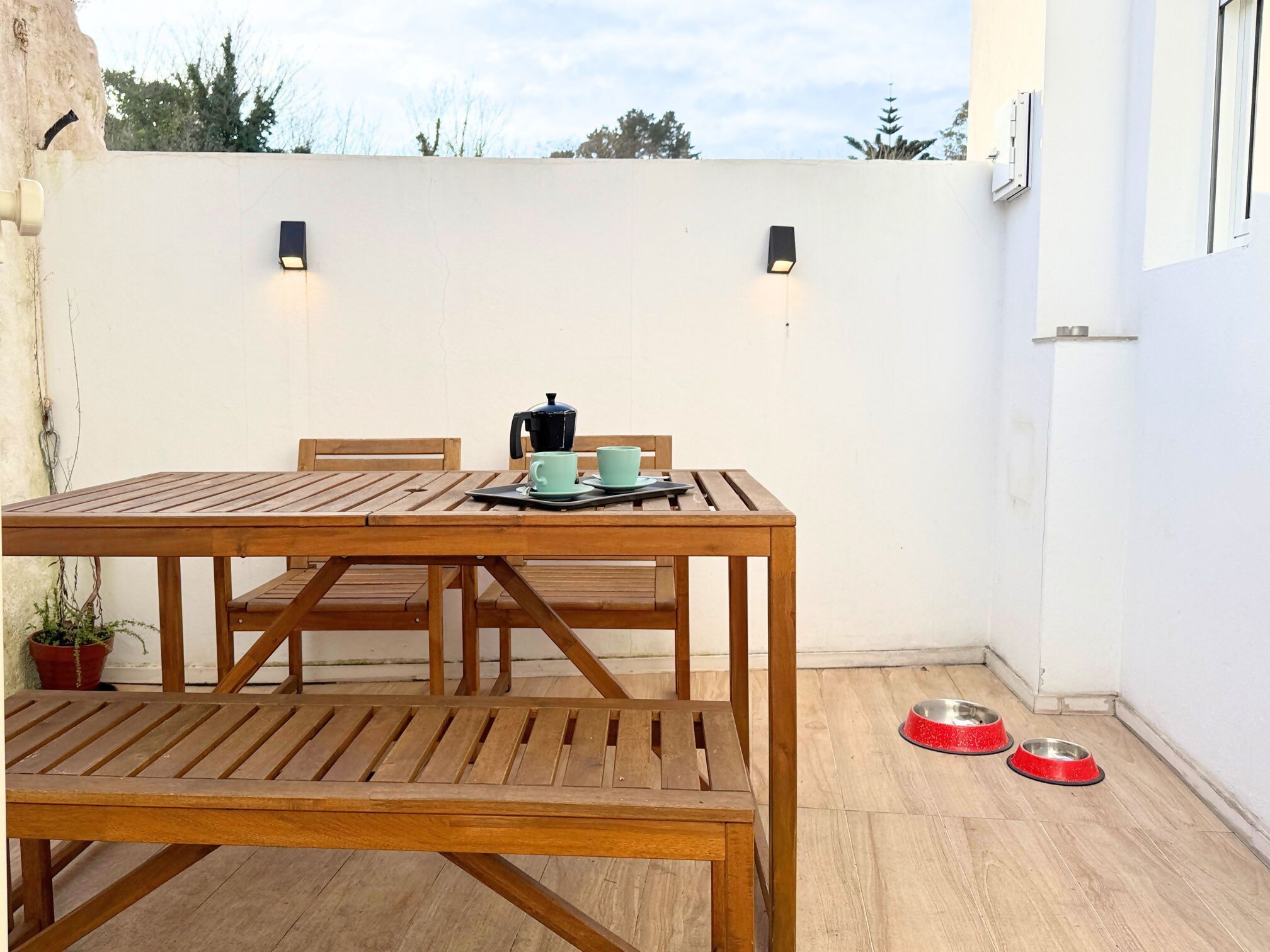 A wooden table with chairs, a coffee pot, and two cups on a patio with a white wall and two wall-mounted lights.
