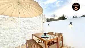 Ein Holztisch mit Stühlen und einem beigen Sonnenschirm auf einer Terrasse mit weißen Wänden und einer weißen Decke.
