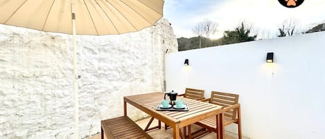 A wooden table with chairs and a beige umbrella in an outdoor patio area with white walls and a white ceiling.
