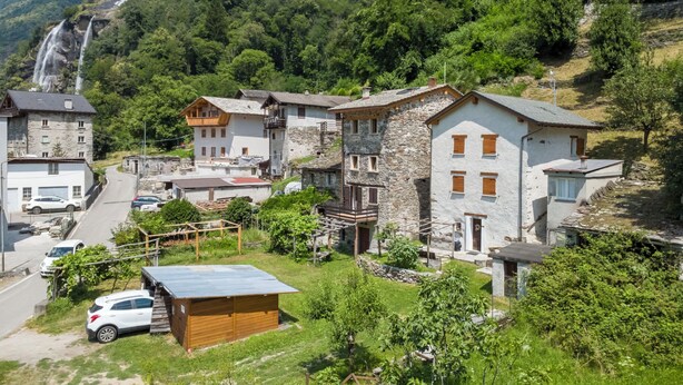 Un village de montagne avec des bâtiments en pierre, une route, des voitures garées et une cascade en arrière-plan.
