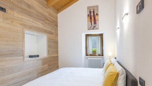 Une chambre avec un mur en bois, un lit avec une literie blanche et jaune, une fenêtre avec des rideaux, un radiateur et un tableau au-dessus de la fenêtre.