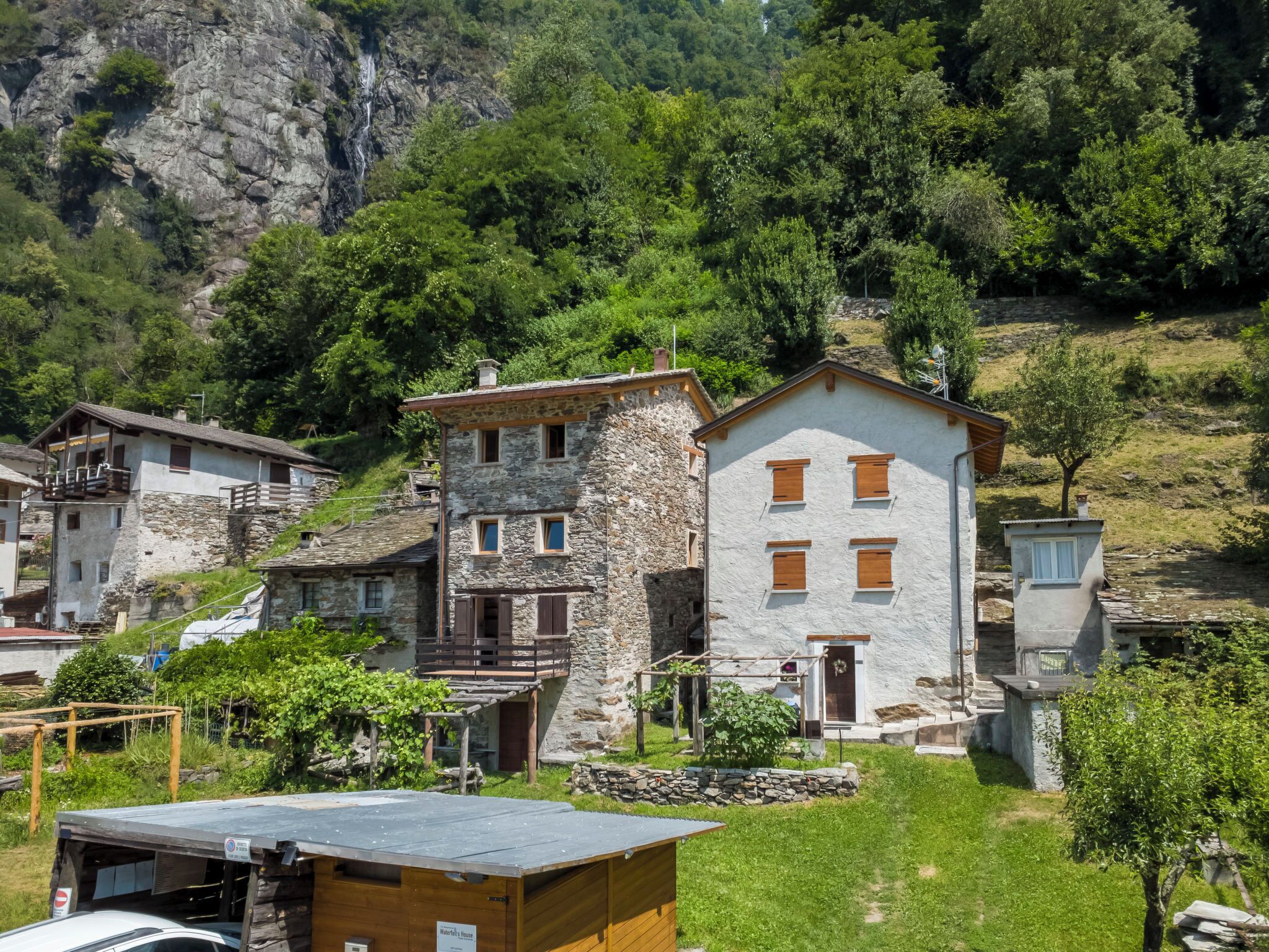 A stone building with multiple windows and a small porch, surrounded by greenery and a mountainous backdrop.