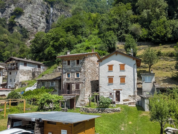 A stone building with multiple windows and a small porch, surrounded by greenery and a mountainous backdrop.