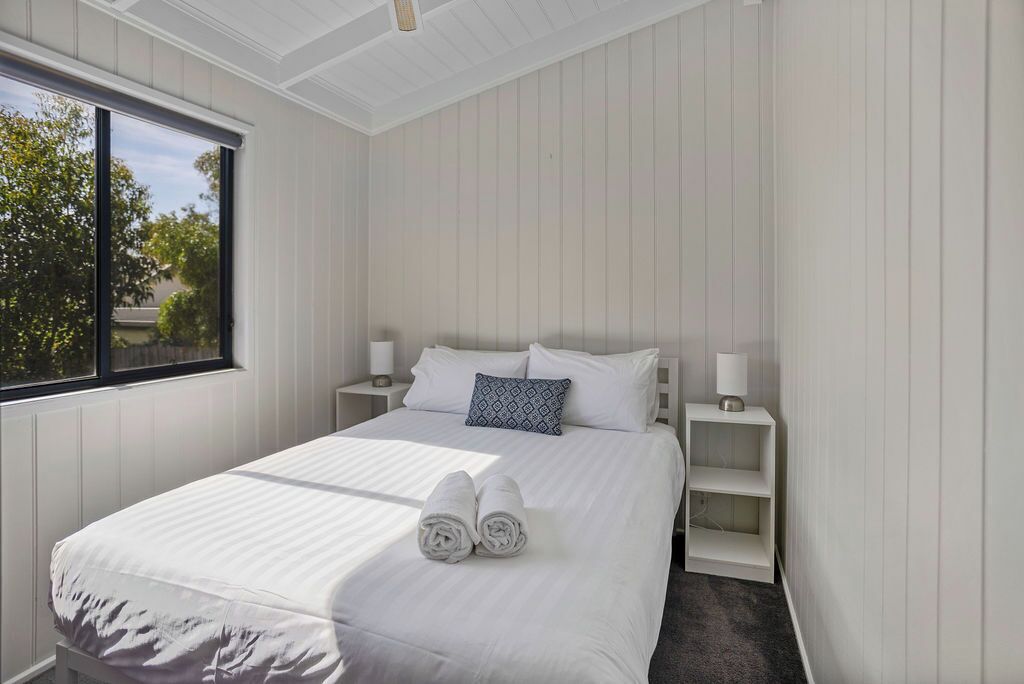 A neatly made bed with white linens and two rolled towels, a window with a view of greenery, and a white wall with a built-in shelf.