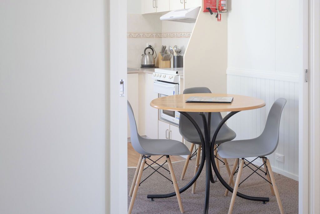 A small dining area with a round table and four chairs, a kitchen in the background with a stove and utensils, and a fire extinguisher on the wall.