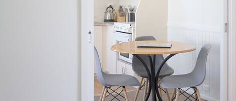 A small dining area with a round table and four chairs, a kitchen in the background with a stove and utensils, and a fire extinguisher on the wall.