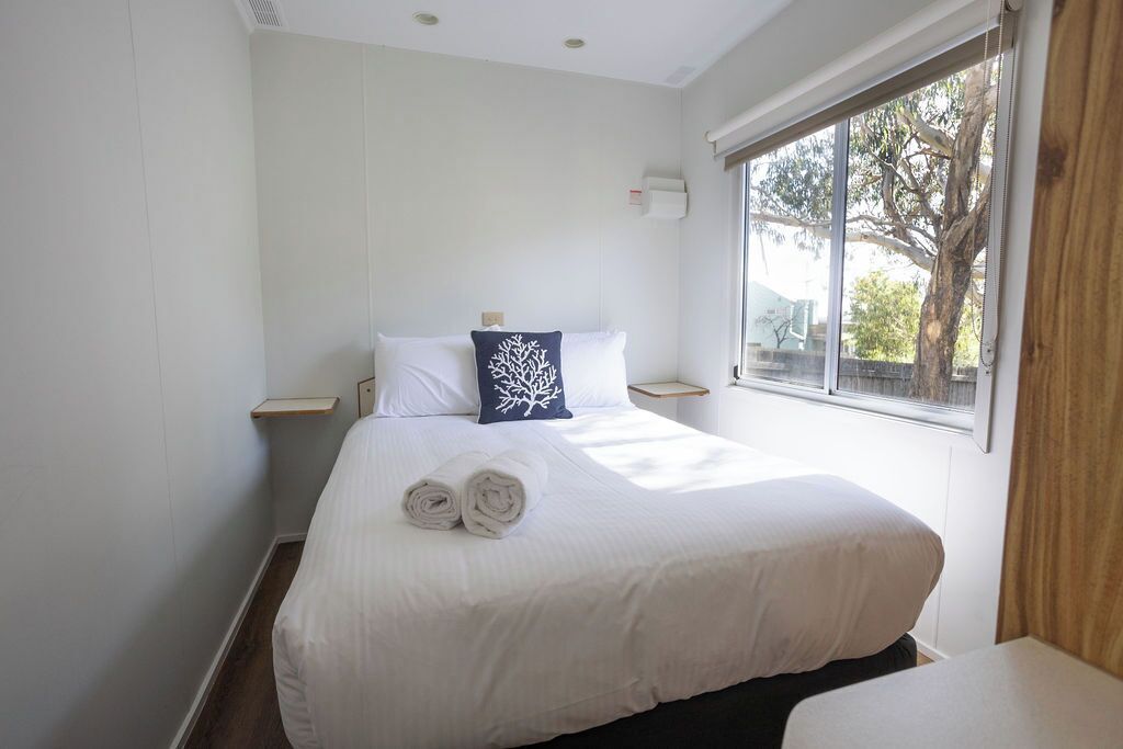 A neatly made bed with white linens and a rolled towel, a nightstand with a lamp, and a window with a view of trees.