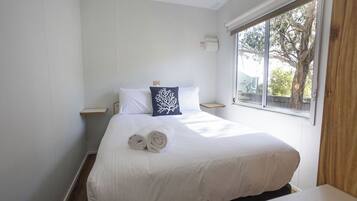 A neatly made bed with white linens and a rolled towel, a nightstand with a lamp, and a window with a view of trees.