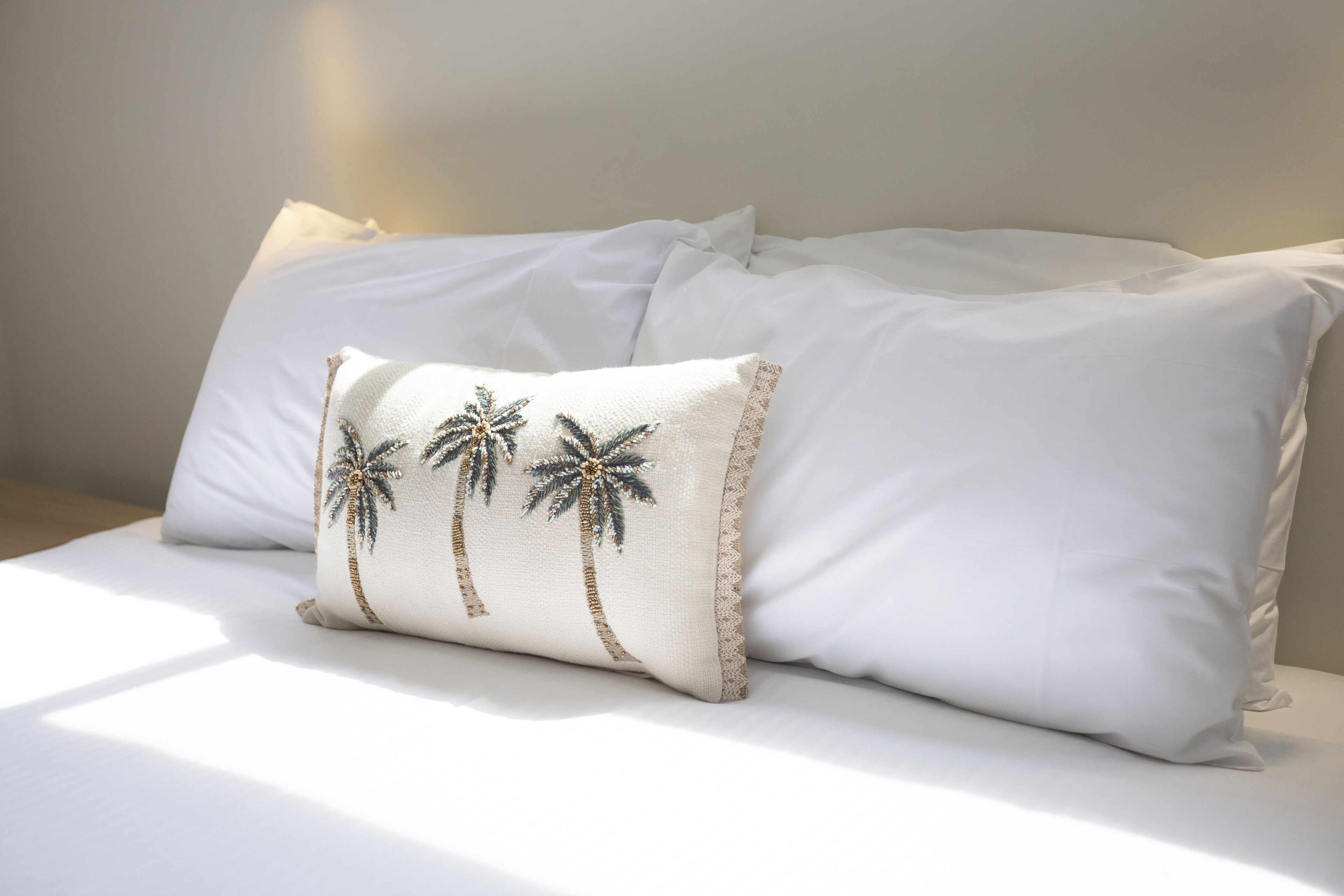 Cama com roupa de cama branca e almofada decorativa com desenhos de palmeiras.