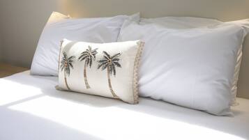 Cama com roupa de cama branca e almofada decorativa com desenhos de palmeiras.