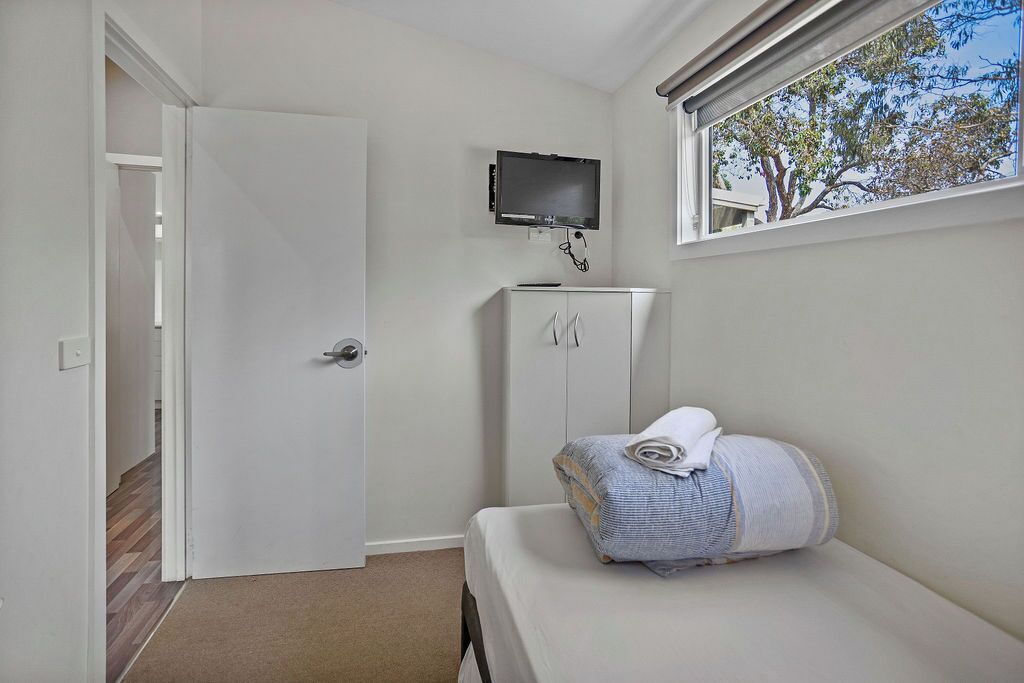Una habitación pequeña y limpia con una cama, un televisor montado en la pared y una ventana con vistas a árboles.