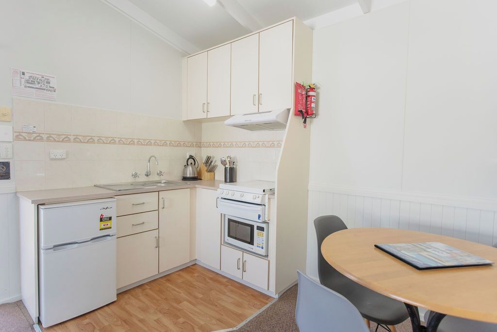 Una cocina compacta con armarios blancos, horno empotrado, microondas y un refrigerador pequeño. Hay una mesa de madera con una revista y una silla.