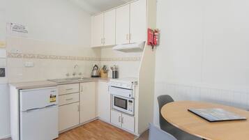 Una cocina compacta con armarios blancos, horno empotrado, microondas y un refrigerador pequeño. Hay una mesa de madera con una revista y una silla.
