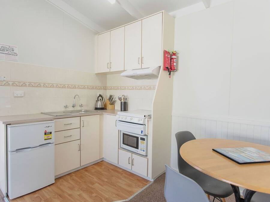 Una cocina compacta con armarios blancos, horno empotrado, microondas y un refrigerador pequeño. Hay una mesa de madera con una revista y una silla.