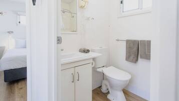 Ein modernes Badezimmer mit Toilette, Waschbecken und Spiegel. Im angrenzenden Raum ist ein Bett zu sehen.