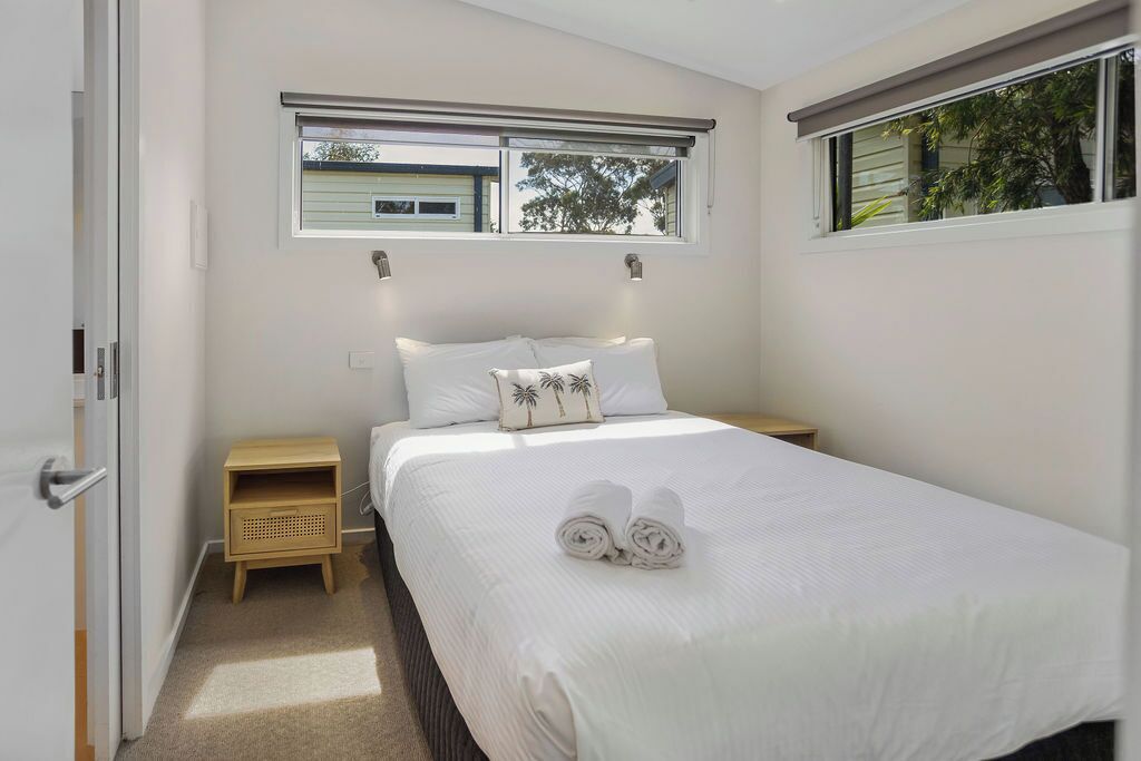A neatly made bed with white linens and two pillows, a small wooden nightstand, a window with blinds, and a door to the left.