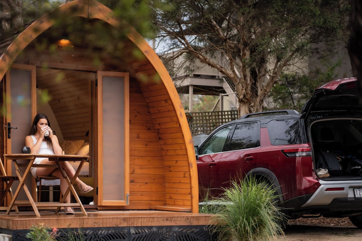 Eine Holzhütte mit Veranda, ein rotes Geländefahrzeug davor geparkt und eine Person, die im Inneren auf einem Stuhl sitzt.