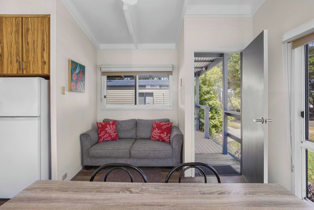 A compact living space with a kitchenette, a grey sofa with red cushions, a dining table, and a view of a deck and greenery outside.
