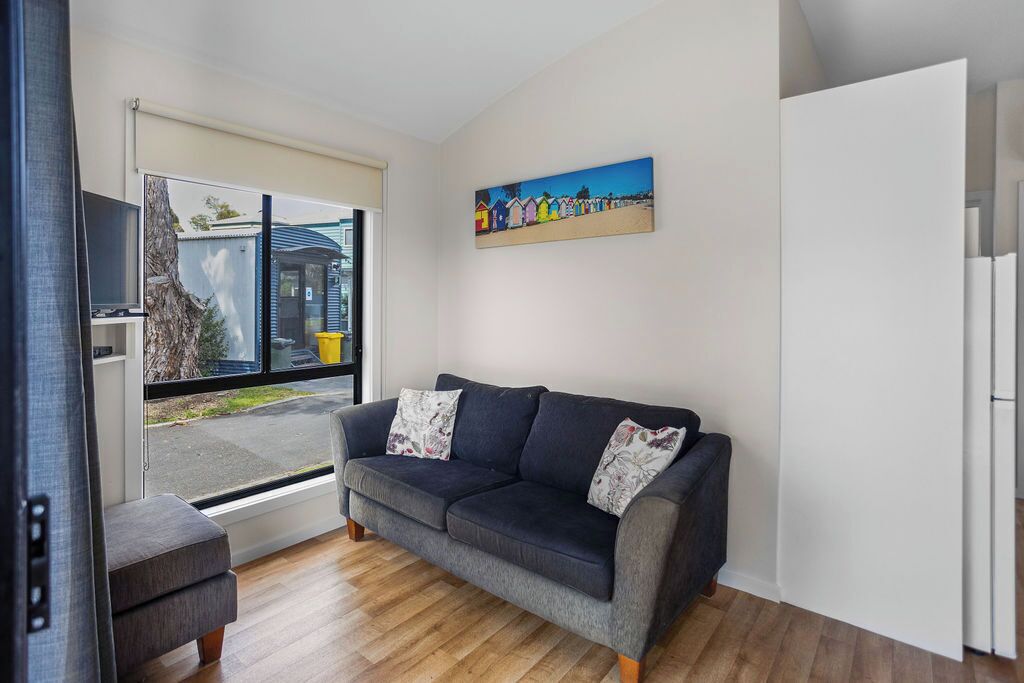 A modern living room with a dark grey sofa, two matching cushions, a small side table, and a large window offering a view of the street and a building outside.