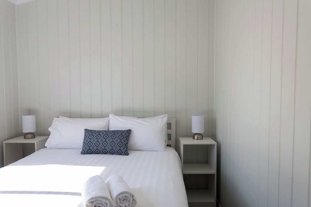 Een netjes opgemaakt bed met witte lakens, twee kussens en een opgerolde handdoek op de zijkant. Er staan twee nachtkastjes met lampen.