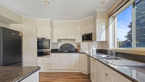 Eine moderne Küche mit weißen Schränken, Granit Arbeitsplatten, einem Edelstahlkühlschrank, zwei Backöfen, einer Mikrowelle und einem großen Fenster mit Blick auf Bäume.
