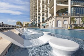 A swimming pool with wave features, a modern building with balconies, and a clear sky. - Luxury 4BR Condo w/Luxury Spa Bathrooms | Lazy River & Resort Amenities (Orange Beach)