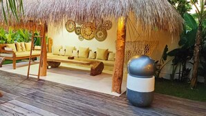 Un espace extérieur aménagé avec une toiture de chaume, comprenant un banc en bois et un fauteuil, le tout entouré d’une végétation luxuriante.