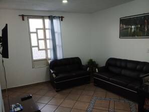 A living room with two black leather sofas, a small table, a potted plant, a painting on the wall, and a window with curtains.