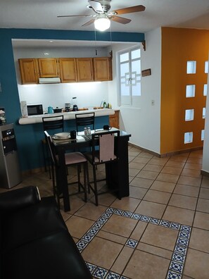 A compact kitchen with wooden cabinets, a dining area with a table and chairs, and a tiled floor with a decorative border.