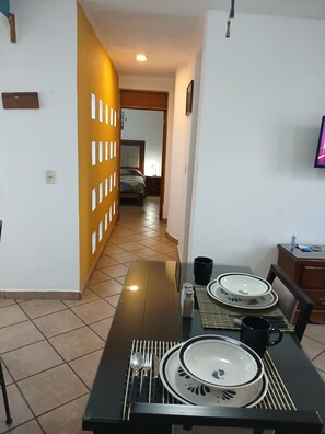 A dining area with a table set for four, a TV mounted on the wall, and a hallway leading to a bedroom.