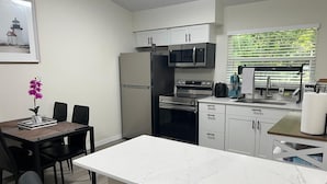 A modern kitchen with white cabinets, a marble countertop, a dining area with a table and chairs, and a window with blinds.