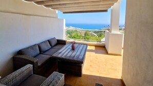 A balcony with a sofa and a table, a view of the sea, and a white wall.
