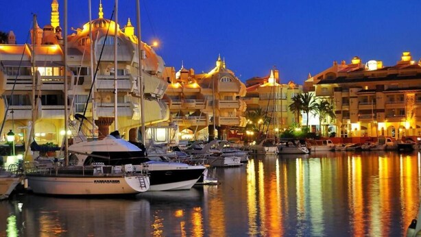 A marina with boats docked, a large building with multiple balconies, and a clear sky at dusk.