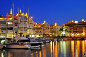 Un puerto deportivo con barcos amarrados, un edificio grande con varios balcones y un cielo despejado al anochecer.