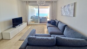 A modern living room with a grey sofa, a flat-screen TV on a white console, a chandelier, and a view of the sea through a sliding glass door.
