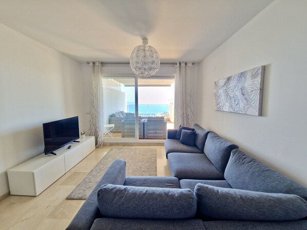 A modern living room with a grey sofa, a flat-screen TV on a white console, a chandelier, and a view of the sea through a sliding glass door.