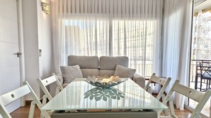 A dining area with a glass table surrounded by white chairs, a sofa with grey cushions, and sheer curtains.