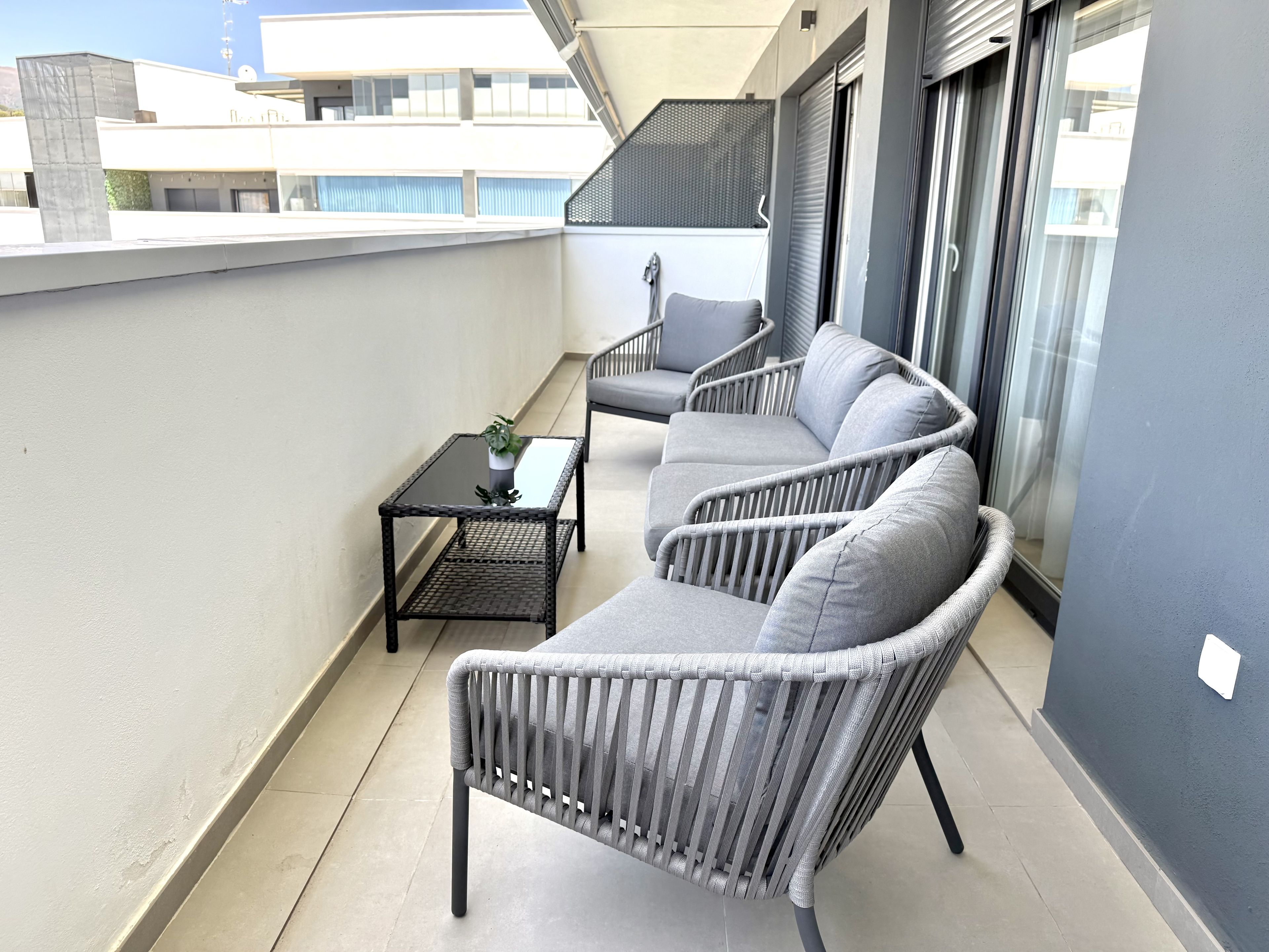 Ein Balkon mit zwei grauen Korbstühlen, einem kleinen Tisch mit einer Pflanze und Blick auf Gebäude.