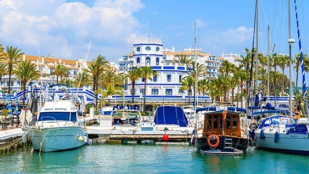 A marina with boats docked, a building with a blue and white facade, and palm trees in the background. - SR Wellingtonia Estepona Downtown (Estepona)