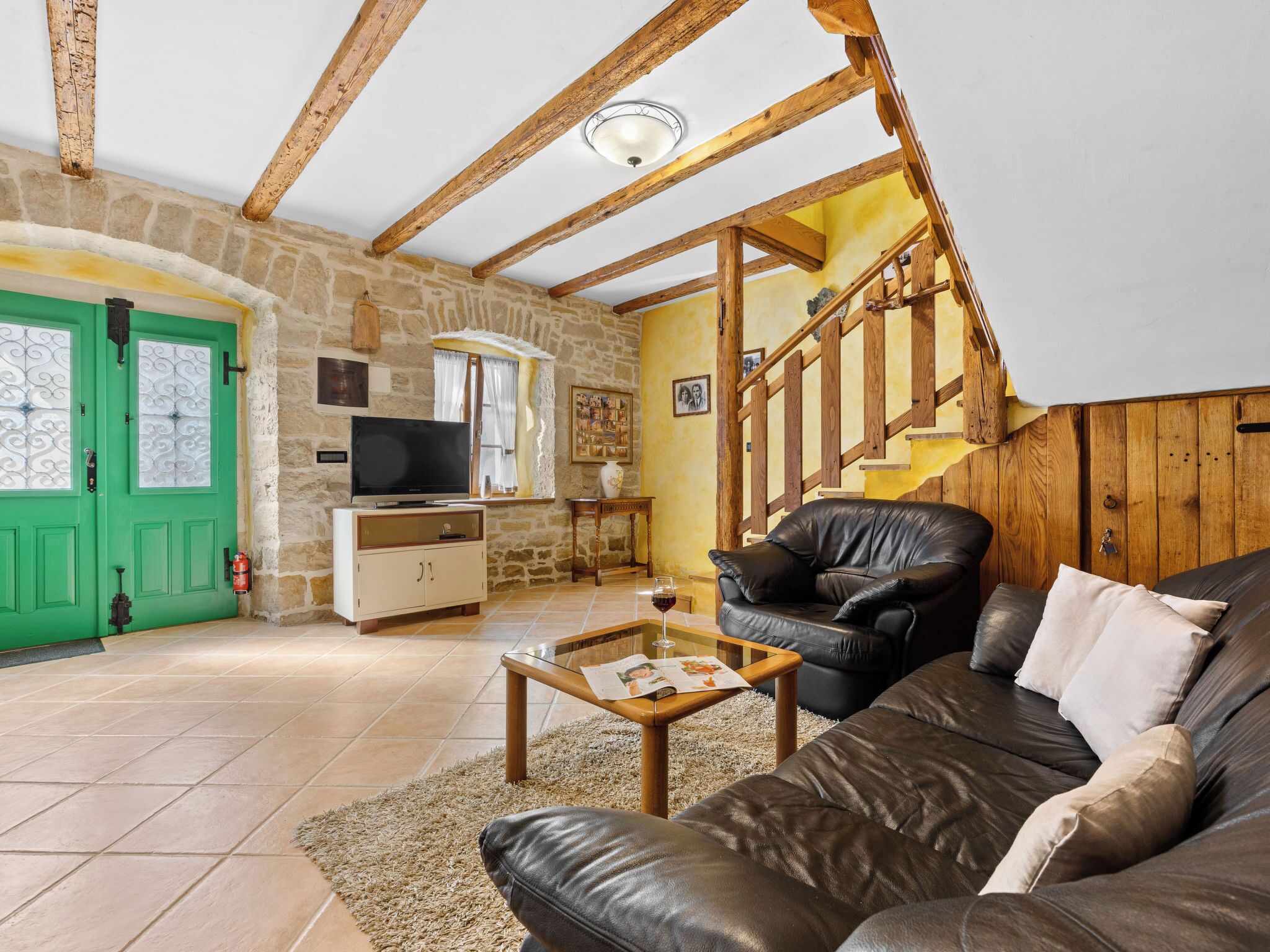 Un salon chaleureux avec un mur en pierre, des poutres en bois, une porte verte, un canapé en cuir, une table basse et un téléviseur.