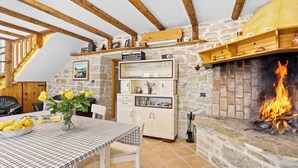 Una cocina acogedora con vigas de madera a la vista, pared de piedra, chimenea encendida y una mesa de comedor con mantel a cuadros.