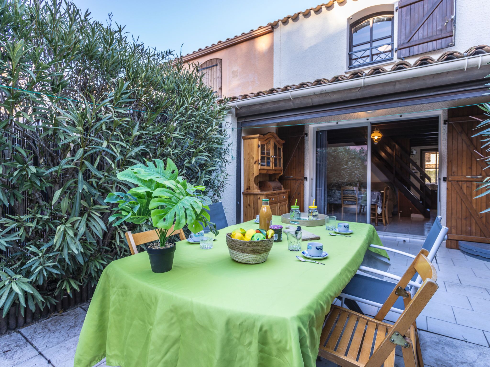 An outdoor dining area with a green table set for two, surrounded by lush greenery and a wooden chair.