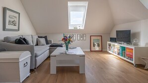 Un loft moderno con techo inclinado, tragaluces, un sofá blanco, una mesa de centro con un jarrón de flores, una estantería y un televisor.