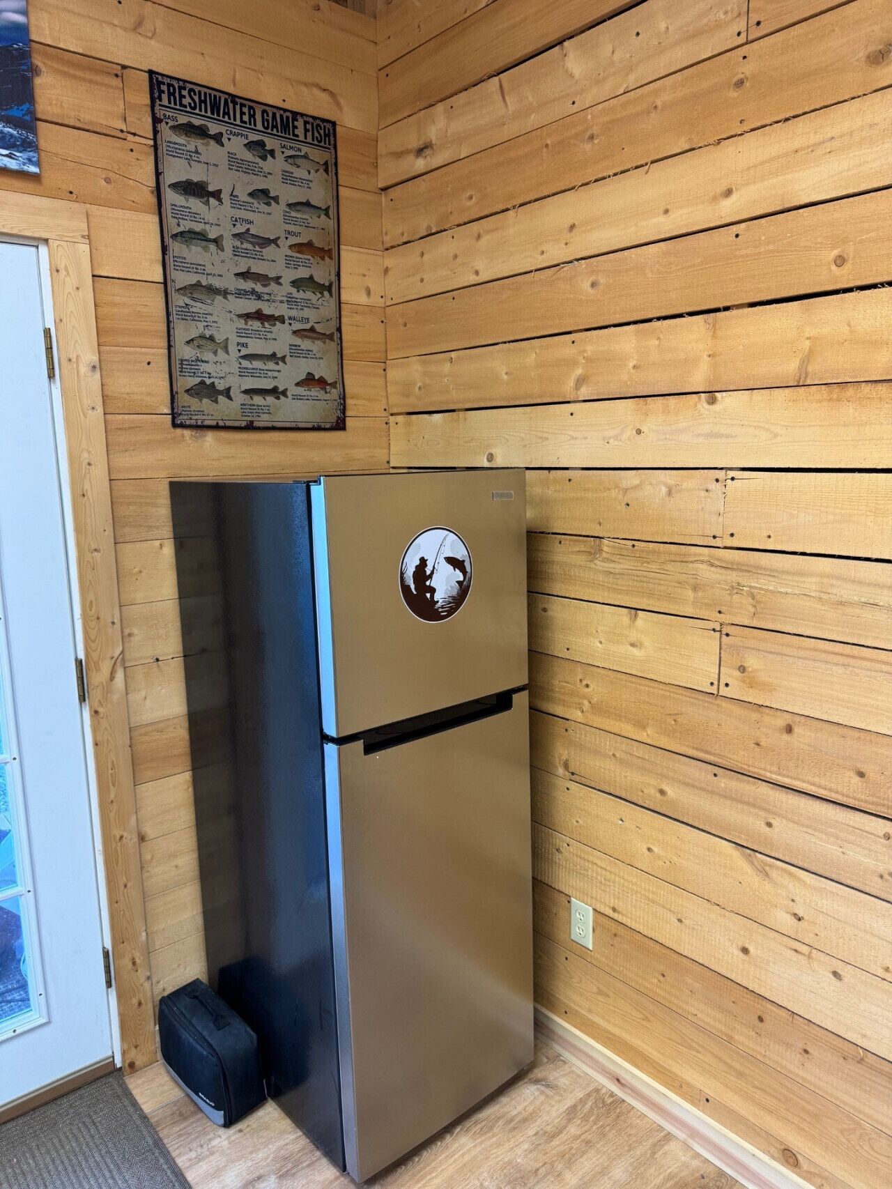 A compact, stainless steel refrigerator with a fishing-themed sticker, positioned against a wooden wall in a room with a door and a poster above it.