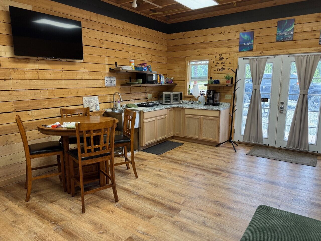 A cozy cabin-style room with a wooden interior, a dining table with chairs, a kitchenette, and a large window with curtains.