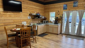 A cozy cabin-style room with a wooden interior, a dining table with chairs, a kitchenette, and a large window with curtains. - Sockeye Chalet at Redbow (Sterling)