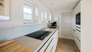 Una cocina moderna con tope de inducción negro, armarios blancos, un lavamanos pequeño y una ventana con persianas.