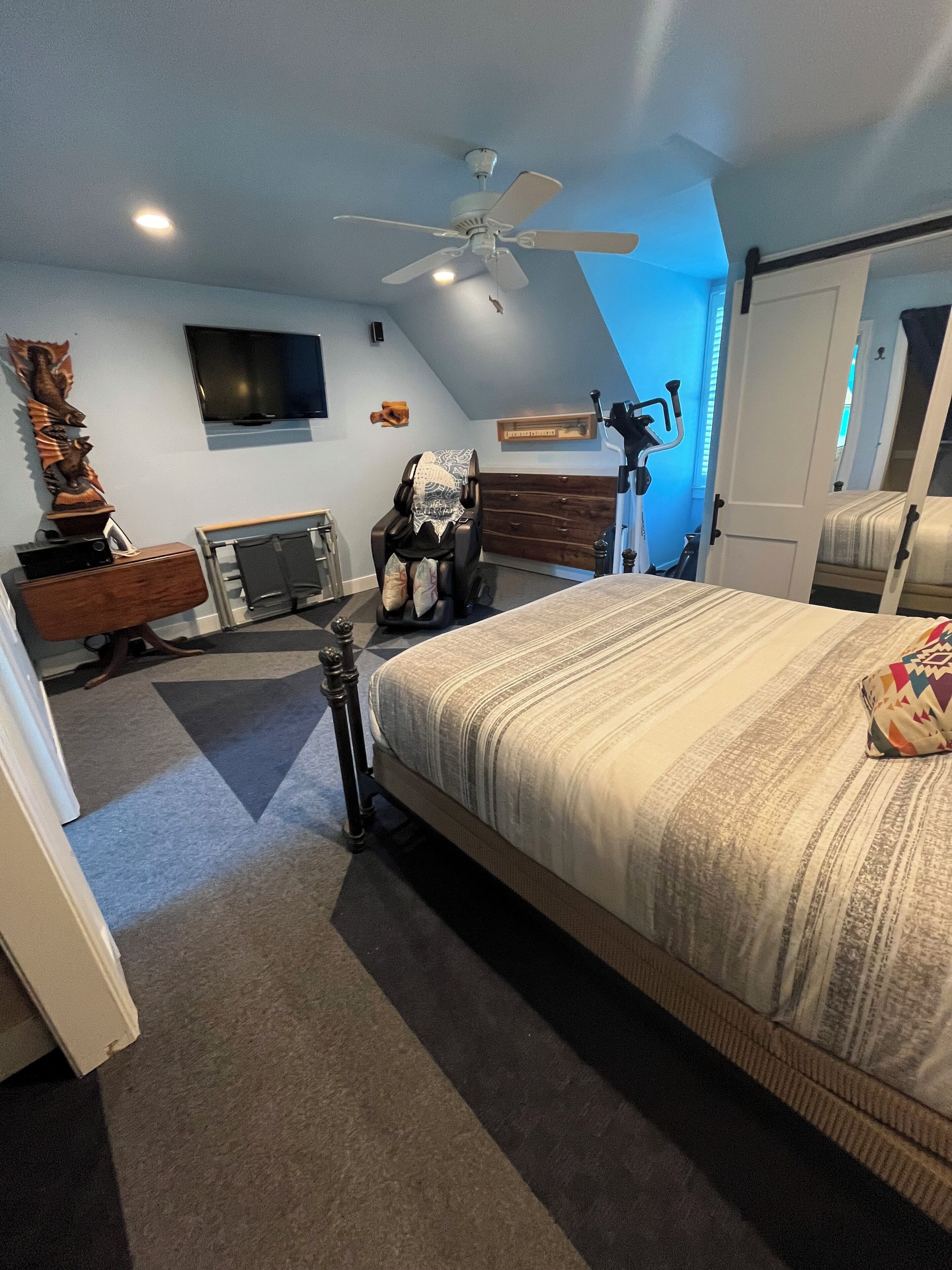 Une chambre à coucher comprenant un lit, un ventilateur de plafond, un fauteuil massant, une télévision et une décoration murale en bois.